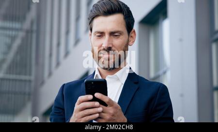 Ein junger, erfolgreicher Geschäftsmann, der sein Smartphone im Geschäftszentrum der Stadt hält, schreibt per Telefon lächelnde Textnachrichten per Handy Stockfoto