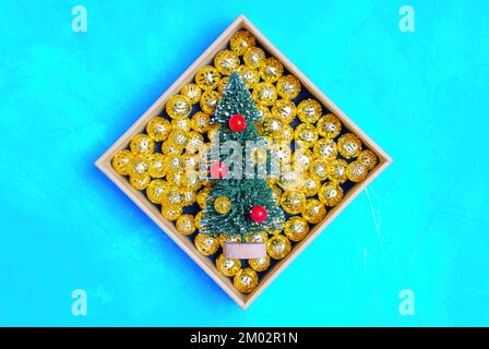 Miniatur-Neujahrsbaum in einer Schachtel mit winzigen goldfarbenen Weihnachtsbällen auf blauem Hintergrund. Stockfoto