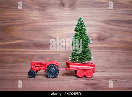 Spielzeugtraktorfigur mit einem kleinen Weihnachtsbaum, isoliert auf dunklem Holzhintergrund in den Wagen gelegt. Neujahrsbaumzucht und -Lieferung. Stockfoto