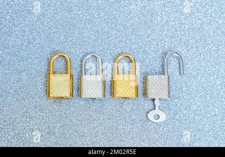 Vier silberfarbene und goldfarbene Vorhängeschlösser auf glitzerndem Hintergrund. Ein Vorhängeschloss wird mit einem Schlüssel geöffnet. Stockfoto