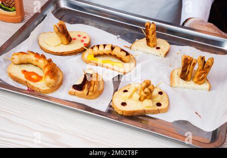 Hot Dog mit verschiedenen Gewürzen und Chili, Paprika und Tomaten und Soßen, Senf und Ketchup auf einem eisernen Tablett auf einem weißen Tisch. Stockfoto