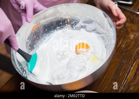 Rühren Sie das Eigelb mit einem Acrylspatel in einer Metallschüssel in das gepeitschte Weiß Stockfoto