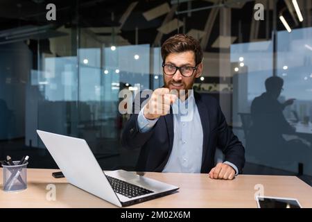 Erfolgreicher, reifer Geschäftsmann, der vor die Kamera schaut und mit dem Finger nach vorne zeigt, um Maßnahmen zu ergreifen, Investor in einen Business-Anzug in einem modernen Büro, der an einem Laptop arbeitet, Chef mit Bart und Brille. Stockfoto
