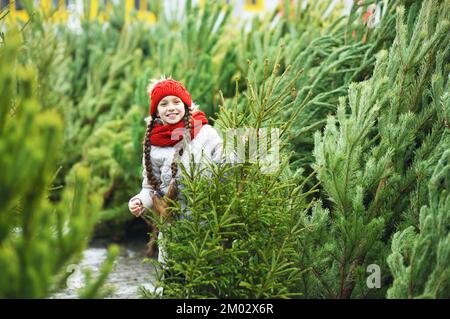 Kleines Mädchen mit Zöpfen in einem roten Strickhut und Schal lächelt auf dem Hintergrund des Weihnachtsbaummarktes und wählt einen Weihnachtsbaum Stockfoto