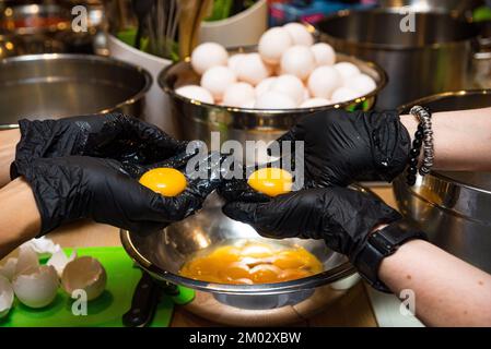 Trennen Sie das Eigelb vom Weiß. Nimm das Eigelb aus der Schüssel. Hände mit schwarzen Handschuhen halten das Eigelb. Ein Haufen Eier im Hintergrund. Stockfoto