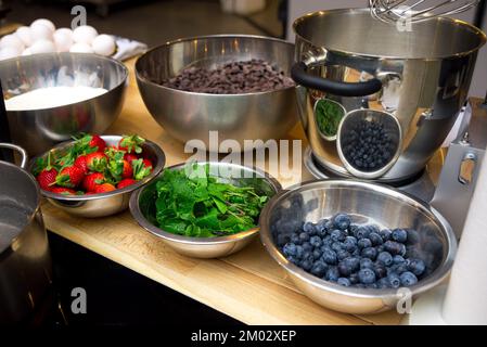 Zutaten für das Dessert in Schüsseln auf dem Tisch. Schokoladentropfen, Mehl, Erdbeeren, frische Minzblätter, Blaubeeren. Stockfoto