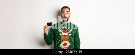Weihnachtsferien und Shopping-Konzept. Überrascht stattlichen Mann in Pullover mit Kreditkarte, stand auf weißem Hintergrund Stockfoto