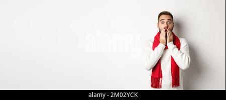 Weihnachtsferien und Feier Konzept. Schöner bärtiger Kerl in rotem Schal und Pullover, der schockiert, beeindruckt von Werbung, weiß Stockfoto