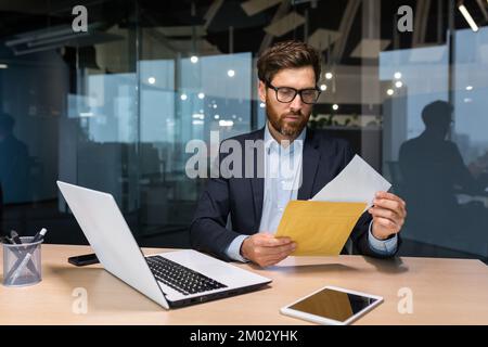 Ein reifer und seriöser Geschäftsmann im Büro, der seinen Brief sorgfältig las, erhielt eine Benachrichtigung, ein Mann im Business-Anzug, der bei der Arbeit ein Notebook benutzte. Stockfoto