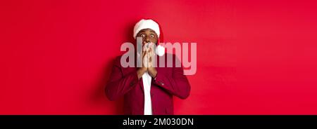 Weihnachts-, Party- und Feiertagskonzept. Schockiert afroamerikanischen Kerl in santa hat Reaktion sprachlos auf Werbung, stand vor rotem Hintergrund Stockfoto