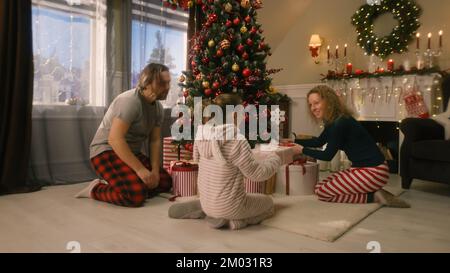 Ein junges Mädchen öffnet Geschenke von Eltern und jubelt am Weihnachtsmorgen unter einem großen, dekorierten Weihnachtsbaum. Warme Atmosphäre zu Hause an Weihnachten oder Neujahr. Glückliche Kindheit. Winterferien. Stockfoto