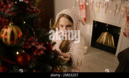 Ein junges Mädchen in einem milden Pyjama kommt zum Weihnachtsbaum, leckt Süßigkeiten und korrigiert Bälle und Spielzeug. Girlande glitzert an der Wand. Vorbereitung vor den Winterferien. Der Zauber des neuen Jahres. Zeitlupe. Stockfoto