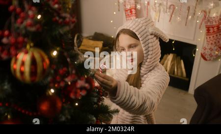 Ein junges Mädchen in einem milden Pyjama kommt zum Weihnachtsbaum, leckt Süßigkeiten und korrigiert Bälle und Spielzeug. Girlande glitzert an der Wand. Vorbereitung vor den Winterferien. Der Zauber des neuen Jahres. Zeitlupe. Stockfoto