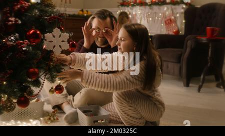 Papa sitzt mit der Tochter im Wohnzimmer und spielt mit Bäumen und Spielzeug. Ein junges Mädchen in einem milden Pyjama schmückt den Weihnachtsbaum. Familienvorbereitung vor Weihnachten oder Neujahr. Stockfoto