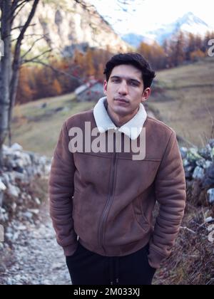 Attraktiver junger Mann in den Bergen im Herbst oder Winter, kaltes Wetter, trägt Wildlederjacke, schaut in die Kamera Stockfoto