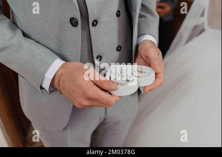 Ein Paar goldene Eheringe in einer weißen Holzkiste in der Form eines Herzens in den Händen eines Mannes. Stockfoto