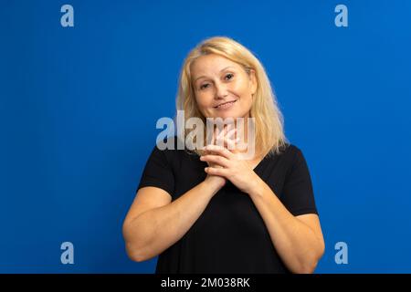 Fotoporträt einer süßen weißen Frau, die ihre Hände zusammenhält und von Bewunderung beeindruckt ist, gekleidet in einem schwarzen T-Shirt. Isoliert auf blauem Studio Stockfoto