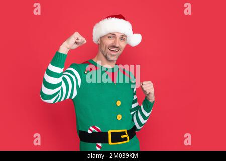 Ein glücklicher erfolgreicher Mann im Elfenkostüm. der weihnachtsmann mit weihnachtsmann-Hut auf rotem Hintergrund. Frohes neues Jahr. Frohe Weihnachten. Stockfoto