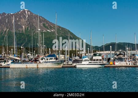 Seward, Alaska, USA - 22. Juli 2011: Alte und neue kleine Boote im Hafen auf blauem Wasser unter blauem Himmel. Bergkette mit Schneeflächen und bewaldetem Grün Stockfoto