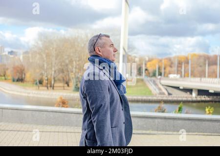 Seitenansicht eines ernsthaften Mannes mittleren Alters mit kurzen Haaren in grauer Jacke, blauem Schal, der auf Fliesen in der Nähe des Flusses steht. Stockfoto