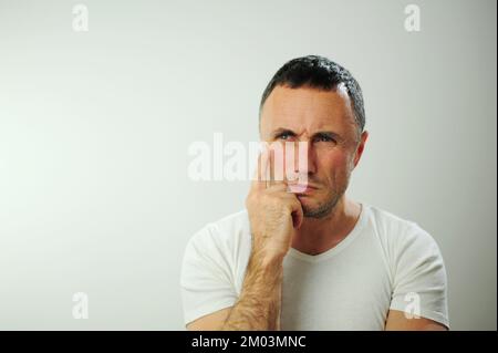 Probleme mit einer Entscheidung strenger, unrasierter Mann schaut in den Rahmen, halte dein Kinn mit der Hand und denke, er hat ein starkes Piercing blaue Augen Er trägt ein weißes T-Shirt mit weißem Hintergrund Stockfoto