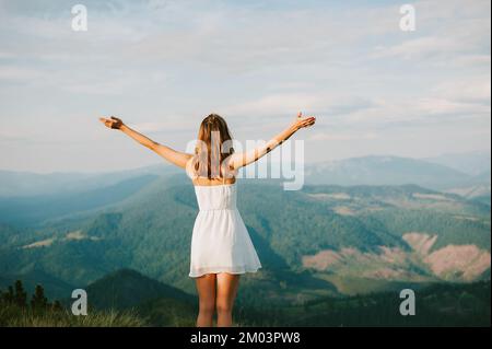 Junge Frau erreicht Berggipfel ein weißes Kleid steht auf Gipfelarmen gestreckt für Erfolg und Freiheit, Rückansicht, Fotoaufnahme in Karpaten Stockfoto