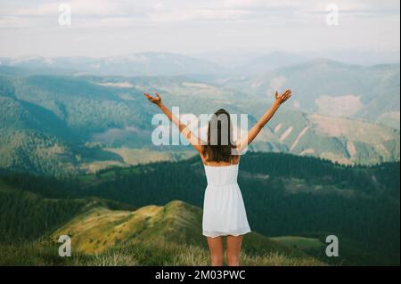 Junge Frau erreicht Berggipfel ein weißes Kleid steht auf Gipfelarmen gestreckt für Erfolg und Freiheit, Rückansicht, Fotoaufnahme in Karpaten Stockfoto