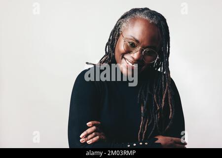 Glückliche professionelle Frau, die in einem Studio eine Brille trägt und dabei die Kamera anlächelt. Eine Frau mittleren Alters mit Dreadlocks, die gegen einen grauen Rücken stehen Stockfoto