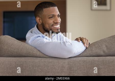 Ein fröhlicher junger afroamerikanischer Mann ruht sich auf einer bequemen Couch aus Stockfoto
