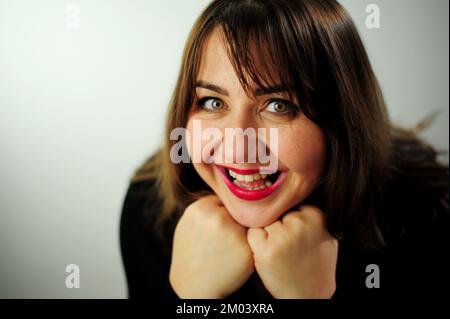 Eine junge Frau lacht auf ihrem Gesicht, ihr Lächeln und ihre Augen leuchten Erwachsene kurvenreiche wunderschöne Frau schaut in den Rahmen Nahgesicht Schwarzes Kleid und roter Lippenstift Sie hat braune Haare Stockfoto