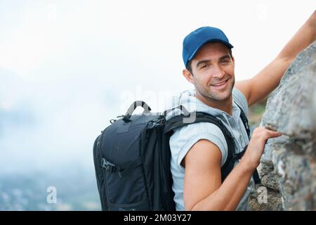 Für diesen Nervenkitzel gibt es keine Grenzen. Junger Kletterer, der die Kamera anlächelt, während er die Klippe hochklettert. Stockfoto