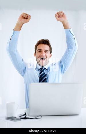 Geschäftssieg. Porträt eines Geschäftsmanns, der an seinem Schreibtisch sitzt, seinen Computer vor sich hat und mit erhobenen Armen über seinem Kopf jubelt. Stockfoto