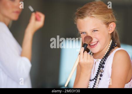 Es kitzelt. Kleines Mädchen probiert Make-up ihrer Mutter an, während ihre Mutter im Hintergrund ist. Stockfoto