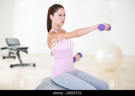 Sie hebt sich in die Fitness. Eine junge Frau, die auf einem Sportball sitzt und ein paar Kraftübungen mit Kurzhanteln macht. Stockfoto