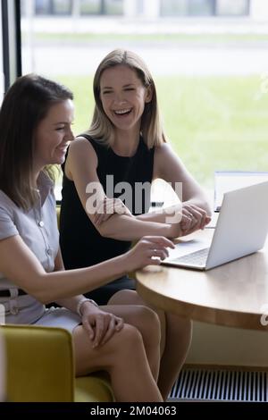 Vertikaler Schuss zwei lachende Frauen sitzen am Tisch mit einem Laptop Stockfoto