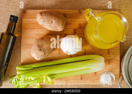 Rezept für Selleriesuppe. Rohes Gemüse wie Selleriestangen, Kartoffeln, Zwiebeln, Olivenöl und Brühe Nahaufnahme auf dem Küchentisch, flach liegend Stockfoto