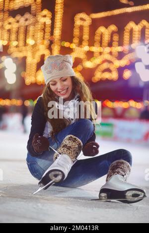 Sie konnte es kaum erwarten, wieder aufzustehen. Eine junge Frau sitzt auf dem Eis einer Eislaufbahn und bindet ihre Schlittschuhe. Stockfoto