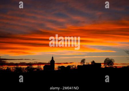 Silhouette eines Dorfes in slowenien, schwarze Silhouette einer Kirche, Häuser und Bäume vor einem wunderschönen roten Sonnenuntergang und majestätischen bewölkten Himmel. Stockfoto