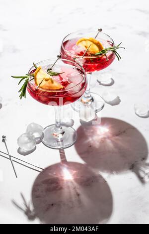 Zwei Weingläser Granatapfel, Eiscocktail mit Rosmarin und Zitrone auf weißem Marmorhintergrund, isoliert mit klaren Schatten in rauem Licht Stockfoto