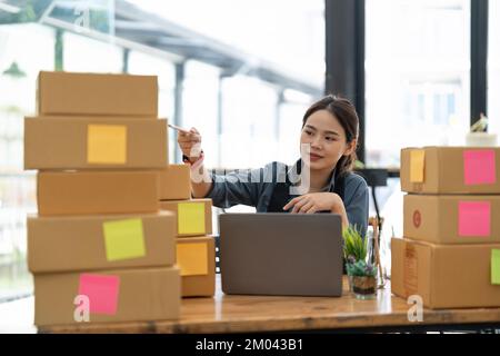 KMU-Unternehmer kleine Unternehmer Online-Verkaufsideen, Happy Young asiatischer Unternehmer arbeitet an Computer und einer Box zu Hause, Lieferung Stockfoto