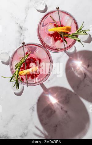 Zwei Weingläser aus Granatapfel, Eiscocktail mit Rosmarin und Zitrone auf weißem Marmorhintergrund, isoliert mit klaren Schatten in rauem Licht Stockfoto