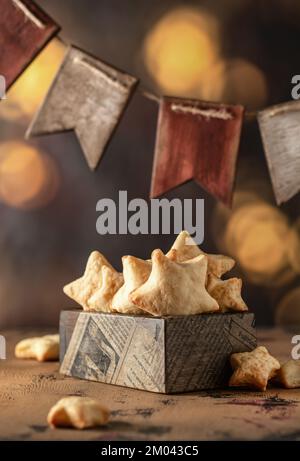 Hausgemachte Zuckerkekse in Sternform mit Zuckerpulver in Holzkiste, mit Girlande aus Holzfahnen und Bokeh-Lichtern im Stockfoto