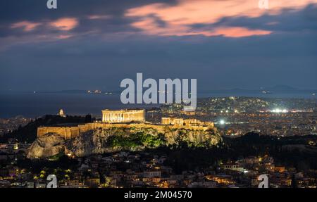 Blick über das Meer der Häuser von Athen, beleuchteter Parthenon-Tempel auf der Akropolis, dramatisch bewölkter Himmel bei Sonnenuntergang, vom Berg Lycabettus, Athen, Gr Stockfoto