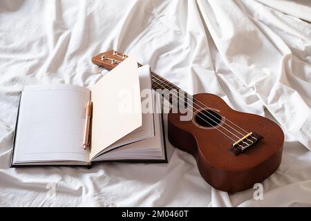 Ukulele-Gitarre und offenes Notizbuch auf weißem Bett. Gemütliche Atmosphäre zum Schreiben von Musik. Stockfoto