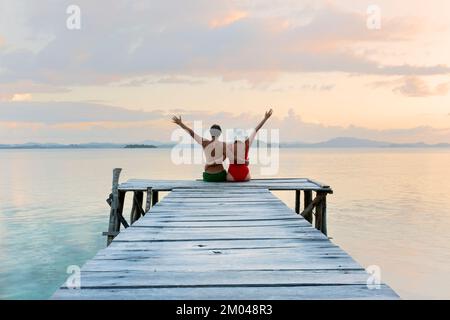 Ein paar Touristen mit den Armen am Himmel sitzen auf dem hölzernen Steg und beobachten den Sonnenuntergang - reisen in Liebe Stockfoto
