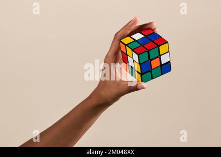 Die Hand einer schwarzen Frau zeigt einen ungelösten Hirn-Teaser des Rubik-Würfels auf beigefarbenem Hintergrund Stockfoto