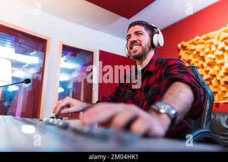 Junger Mann, der in einem professionellen Musikstudio mit einem Computer und Kopfhörern einen Song ausgleicht. Fröhliche Toningenieurin, die arbeitet und das Stück mixt. Technologiekonzept. Hochwertiges Foto Stockfoto