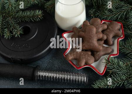 Kurzhanteln, Gewichtsteller, Lebkuchen, Weihnachtskekse, ein Glas Milch für den Weihnachtsmann, Baumzweige. Winter-Fitness-Diät, Fitness-Fitness-Diät-Konzept. Stockfoto