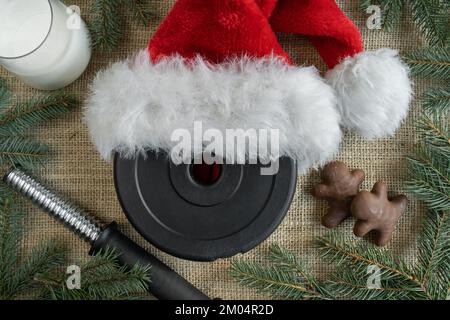 Gym Hantelplatte mit rotem Weihnachtsmann Hut, Weihnachtskekse, Glas Milch, Baumzweige. Winter Fitness Diät, Training Diet Konzept. Stockfoto