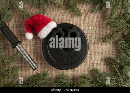 Kurzhantel-Belag mit rotem Weihnachtsmann-Hut. Fitness-Feiertage Winterzusammensetzung. Fitnesskonzept mit Weihnachtsbaumzweigen. Stockfoto